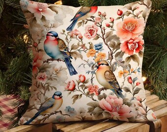 Puede incluir: Cojín decorativo cuadrado con diseño floral y de pájaros. El cojín crema presenta pájaros coloridos posados en ramas con flores rosas, naranjas y azules. El cojín está colocado sobre una superficie de madera.