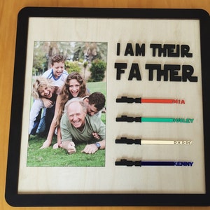 May include: A framed photo of a family with the text "I AM THEIR FATHER" above the photo. Below the photo are four lightsabers with the names Mia, Harley, Bobby, and Kenny written next to them.