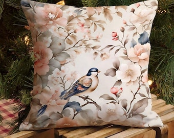 Puede incluir: Cojín decorativo cuadrado con un diseño floral y de pájaros. El cojín presenta un fondo crema con flores rosas y melocotón, hojas grises y un pájaro azul y marrón posado en una rama. El cojín está colocado sobre una superficie de madera.