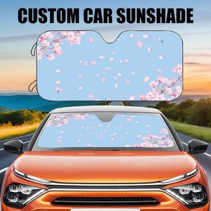 May include: A custom car sunshade with a light blue background and a floral design of pink cherry blossoms and falling petals. The text "CUSTOM CAR SUNSHADE" is displayed at the top. The sunshade is installed in the windshield of an orange car.