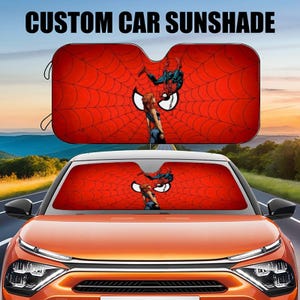 May include: Custom car sunshade featuring a red and black spider web design with Spiderman and a woman in a red and black costume. The sunshade is installed in a car windscreen.