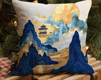 Puede incluir: Cojín decorativo cuadrado con un diseño de paisaje bordado. El cojín tiene un fondo color crema con una escena de montaña en tonos azules, dorados y blancos. También se presenta un gran sol dorado y una pagoda.