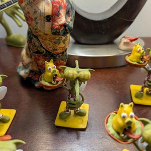 May include: A collection of whimsical figurines, including green alien-like creatures with large eyes and yellow dog-like figures. The figurines are on yellow bases and some are holding small plates. A decorative cat statue is also present.
