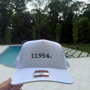 May include: White baseball cap with the number "11954." embroidered on the front. The cap has a curved brim and a tag that reads "OTTO Classic." The background features a pool and trees.
