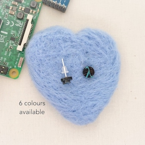 Puede incluir: Una bola de lana azul en forma de corazón con dos pequeños botones negros y verdes encima. El texto "6 colores disponibles" está impreso debajo de la lana.