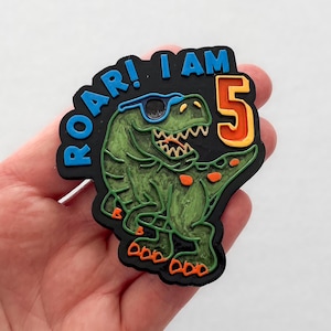 Puede incluir: Accesorio con temática de dinosaurio con el texto "ROAR! I AM 5". El dinosaurio es verde con manchas naranjas y gafas de sol. El número 5 es naranja con contorno amarillo. Ideal para un quinto cumpleaños.