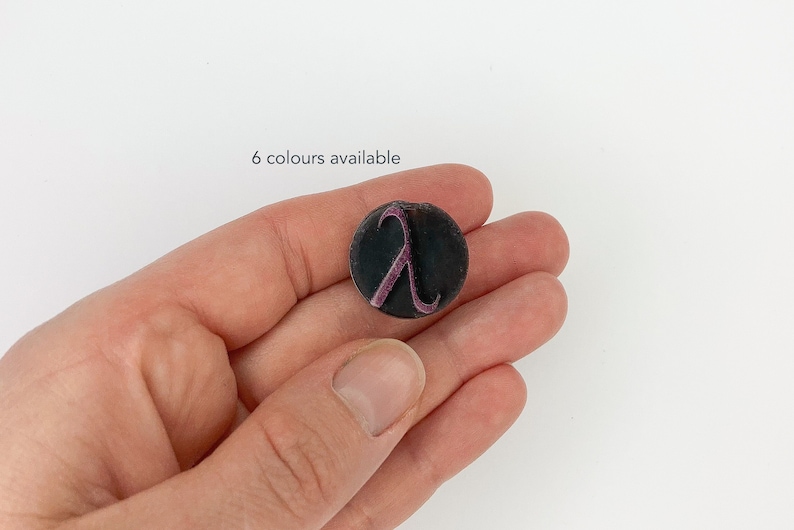 May include: A circular, dark grey button featuring a purple lambda symbol. The text "6 colours available" is above the button. The button is held in a hand against a white backdrop.