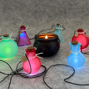 Puede incluir: Una colección de botellas de pociones brillantes en varios colores, incluyendo verde, rosa y azul, con tapones de corcho y cuerdas negras adjuntas. Un caldero negro con una vela encendida se encuentra en el centro. Las botellas están dispuestas sobre una superficie gris.