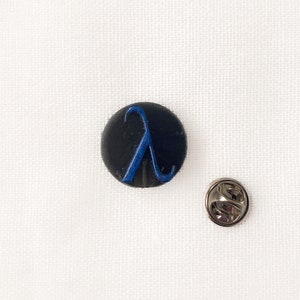 May include: A black, circular pin featuring a blue lambda symbol. Beside it is a silver-coloured pin back. The items rest on a white, textured surface. This pin is a great accessory for jackets, bags, or hats.