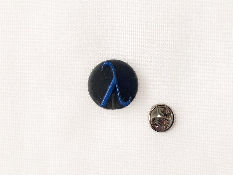 May include: A black, circular pin featuring a blue lambda symbol. Beside it is a silver-coloured pin back. The items rest on a white, textured surface. This pin is a great accessory for jackets, bags, or hats.