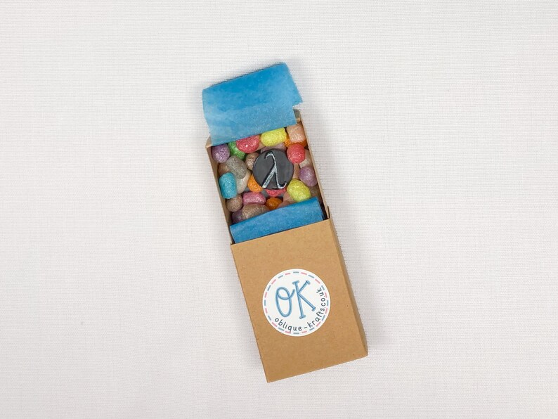 May include: A small, open cardboard box filled with colourful, pebble-shaped sweets and a black charm. The box is lined with blue tissue paper and has a circular sticker with the text "OK oblique-krafts.co".