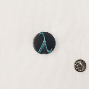 May include: A black circular badge featuring a teal lambda symbol. The badge is set against a white backdrop, with a silver pin back. A small accessory.
