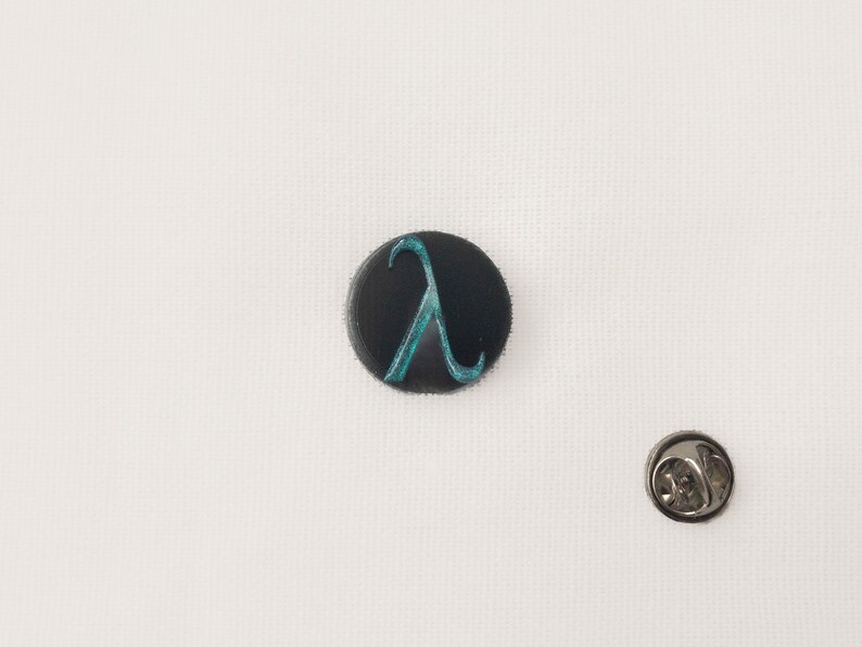 May include: A black circular badge featuring a teal lambda symbol. The badge is set against a white backdrop, with a silver pin back. A small accessory.