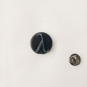 May include: A black, circular badge featuring a silver Lambda symbol, alongside a separate silver pin. The items are set against a white backdrop.