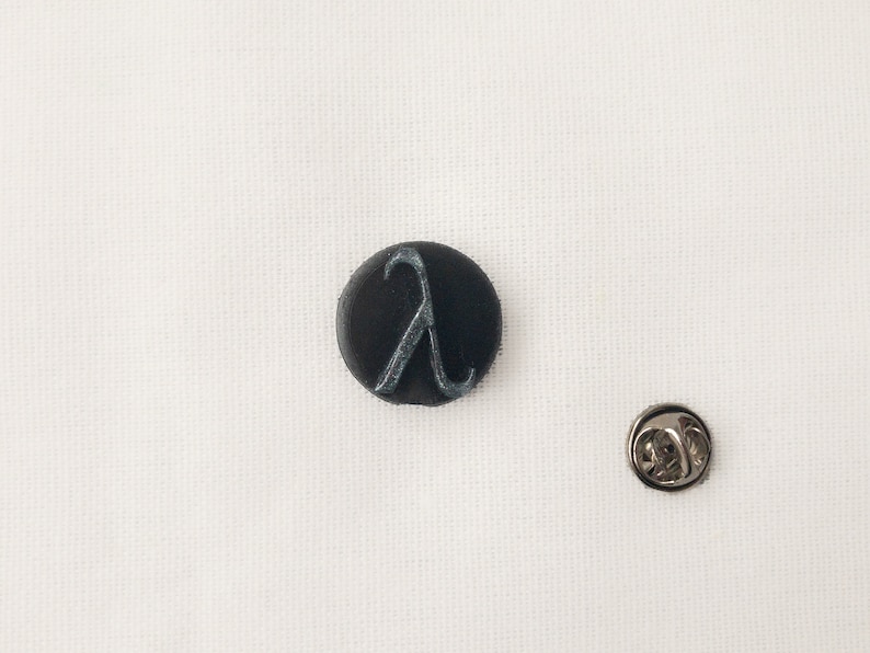 May include: A black, circular badge featuring a silver Lambda symbol, alongside a separate silver pin. The items are set against a white backdrop.