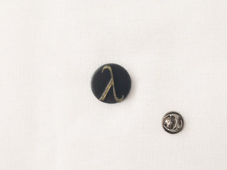 May include: A black, circular pin featuring a gold lambda symbol. A silver pin back is positioned nearby. The pin is set against a white backdrop.