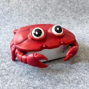 Puede incluir: Un recipiente rojo con forma de cangrejo, con grandes ojos de dibujos animados. El recipiente tiene una base blanca y pinzas rojas. El diseño es caprichoso y juguetón, adecuado para guardar objetos pequeños.