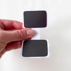 May include: A white paper sign with two black rectangular areas, connected by a white stem. The sign is held by a hand against a white background. The edges of the sign are uneven.