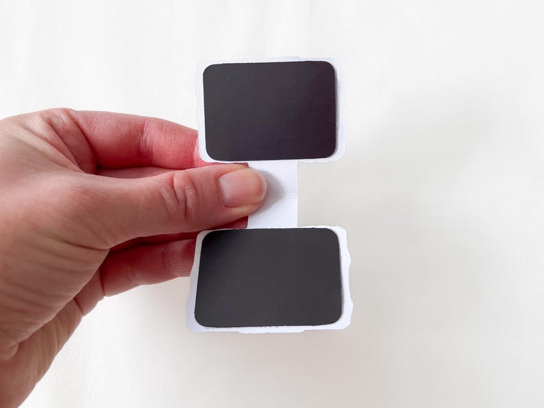 May include: A white paper sign with two black rectangular areas, connected by a white stem. The sign is held by a hand against a white background. The edges of the sign are uneven.