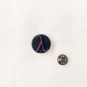 May include: A black circular badge featuring a purple lambda symbol. A silver pin back is positioned beside the badge on a white fabric. This is likely a lapel pin or a decorative badge.