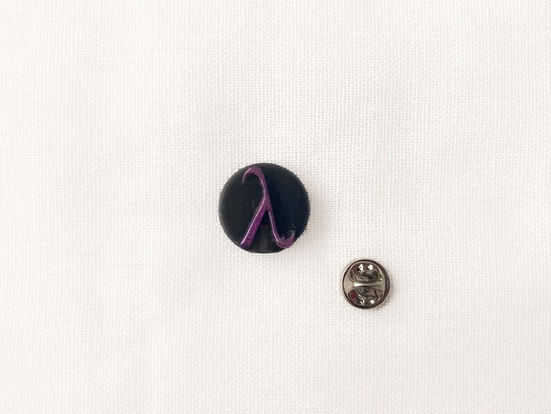 May include: A black circular badge featuring a purple lambda symbol. A silver pin back is positioned beside the badge on a white fabric. This is likely a lapel pin or a decorative badge.