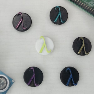 May include: A selection of circular buttons with the Greek letter lambda. The buttons are in various colours, including black, white, purple, blue, and green. The lambda symbol is raised and metallic.