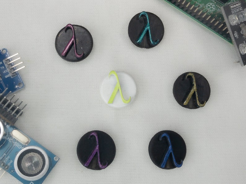 May include: A selection of circular buttons with the Greek letter lambda. The buttons are in various colours, including black, white, purple, blue, and green. The lambda symbol is raised and metallic.