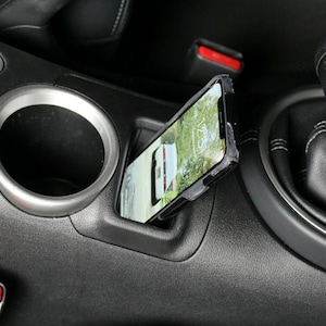 May include: A black phone case with a silver trim is inserted into a car's center console. The phone screen shows a map with a white car icon. The car's gear shift is visible in the background.