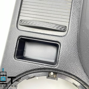 May include: Black plastic car interior trim piece with a rectangular storage compartment and a small, rectangular, carbon fiber-look handle.