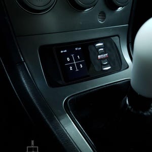 May include: A black and white digital display with the text "air-out" and "137 psi" on the screen. The display is mounted in a car's dashboard and has four buttons labeled "0", "1", "2", and "3". The display is next to a black button with the text "AIRLIFT" in white.