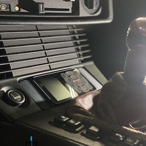 May include: Close-up of a car's interior with a black leather gear shift knob, a black dashboard with a digital display, and a black and silver control panel with buttons. The display shows the text "AIRLIFT" in red.