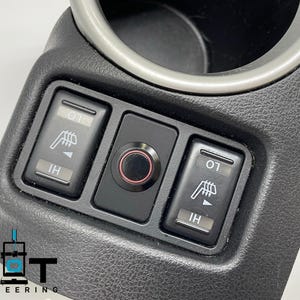 May include: Close-up of a black car interior with three buttons. The buttons are labeled "O1" and "IH" and have a graphic of a seat with arrows. The button in the middle is a round, black button with a red ring around it.
