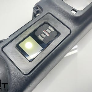 May include: A black plastic panel with a rectangular cutout for a screen. The screen is illuminated with a white light. The panel has a small black button with the word "SHIFT" printed on it.