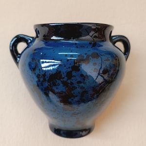 Könnte beinhalten: Eine kleine, blaue Keramikvase mit schwarzen Flecken und zwei Henkeln. Die Vase hat eine glänzende Oberfläche.