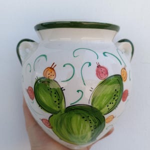 Op de afbeelding: Een witte keramische vaas met twee kleine handvatten en een groene rand. De vaas is versierd met groene cactusvormen, oranje en roze fruit en groene krullen. De vaas wordt in een hand gehouden.