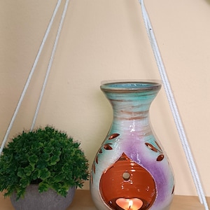 Puede incluir: Un difusor de aceites esenciales de cerámica decorativo con una vela encendida en su interior. El difusor en forma de jarrón tiene cortes en forma de hoja y está pintado con tonos de verde azulado, morado y marrón. Una pequeña planta en maceta está a la izquierda.