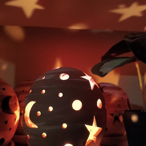 Puede incluir: Un portavelas esférico de cerámica oscura con recortes de estrellas, lunas y puntos. La luz brilla a través de los recortes, proyectando formas de estrellas en el fondo. El portavelas se encuentra sobre un platillo.