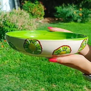 Puede incluir: Un cuenco de cerámica con interior verde brillante y exterior color crema. El cuenco está decorado con rodajas de kiwi. El cuenco se sostiene frente a un fondo verde y herboso.
