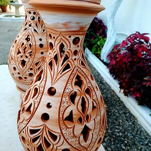 May include: Two terracotta-colored decorative vases with intricate cut-out patterns. The vases have a bulbous shape with a flared base and a rounded top. The tops are painted white. The background includes greenery and a white wall.