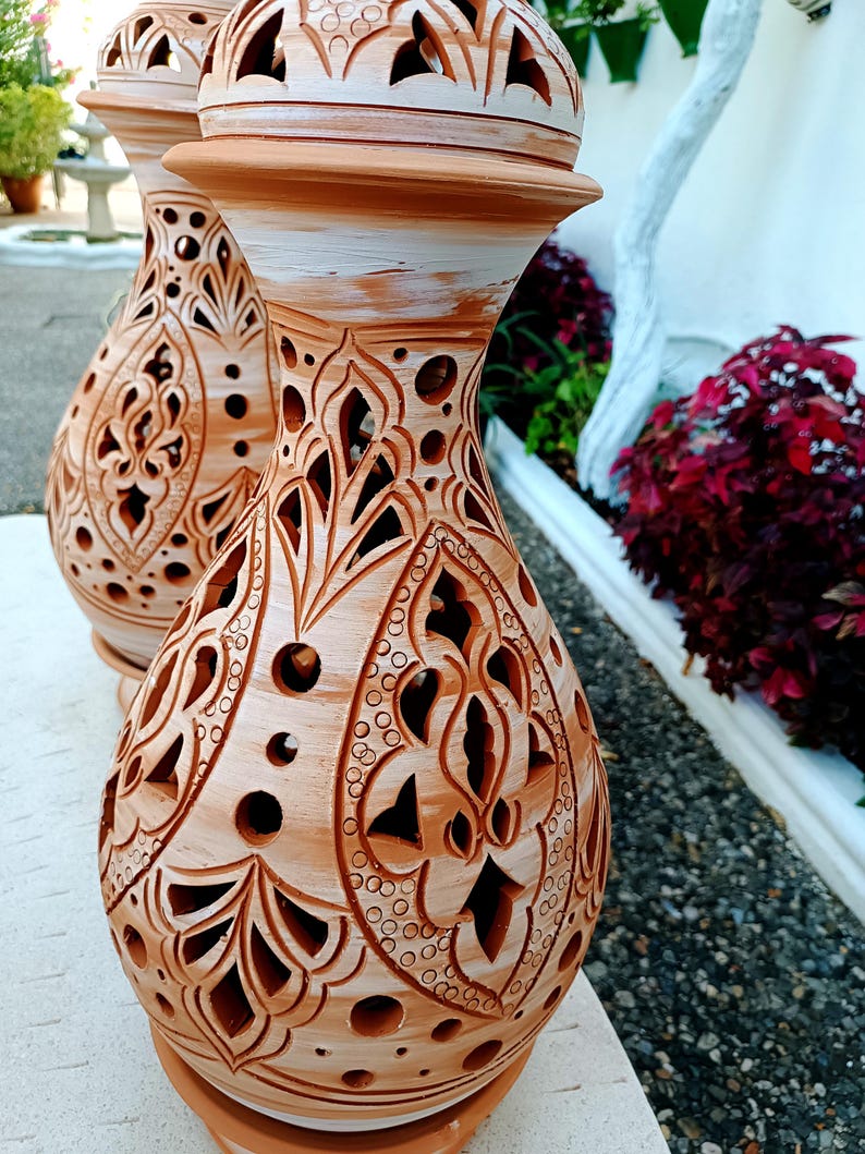 May include: Two terracotta-colored decorative vases with intricate cut-out patterns. The vases have a bulbous shape with a flared base and a rounded top. The tops are painted white. The background includes greenery and a white wall.