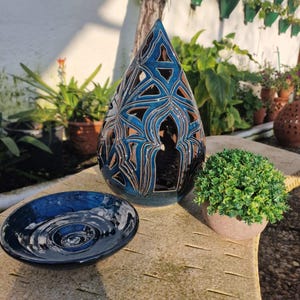 Könnte beinhalten: Ein blauer Kerzenhalter aus Keramik in Tropfenform mit filigranen Ausschnitten. Der Halter steht auf einem Tisch mit einer kleineren blauen Keramikschale und einer Topfpflanze.