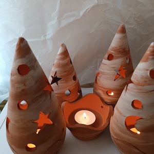 Op de afbeelding: Vier terracotta kaarshouders in de vorm van kerstboom met uitgesneden sterren en cirkels. De kaarshouders zijn verlicht met theelichtjes.