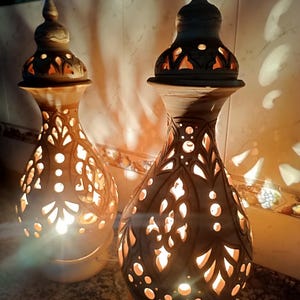 May include: Two ceramic lamps with intricate cut-out patterns, emitting a warm glow. The lamps have a bulbous body and a pointed top, showcasing a traditional design. The lamps are beige and brown, with a light source inside.