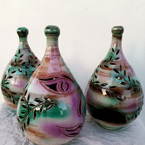 Può includere: Tre portacandele in ceramica con una glassa verde, marrone e viola. Ogni portacandele ha un design unico con un viso e foglie ritagliate.
