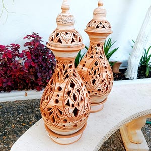 May include: Two terracotta-colored, ornate ceramic vases with intricate cut-out designs. Each vase has a bulbous body, a flared base, and a decorative, pointed top. They sit on a white bench, with greenery in the background.