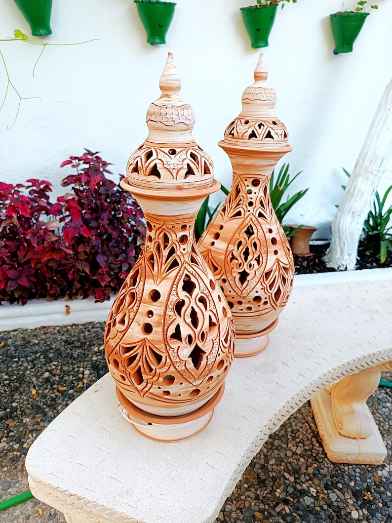 May include: Two terracotta-colored, ornate ceramic vases with intricate cut-out designs. Each vase has a bulbous body, a flared base, and a decorative, pointed top. They sit on a white bench, with greenery in the background.