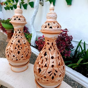 May include: Two terracotta-colored ceramic lanterns with intricate cut-out designs. Each lantern has a bulbous body, a decorative top, and sits on a round base. The lanterns are displayed outdoors on a stone bench, with greenery in the background.