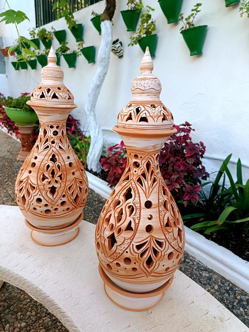 May include: Two terracotta-colored ceramic lanterns with intricate cut-out designs. Each lantern has a bulbous body, a decorative top, and sits on a round base. The lanterns are displayed outdoors on a stone bench, with greenery in the background.