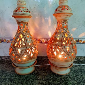 May include: Two decorative ceramic lamps with intricate cut-out patterns, emitting a warm orange glow. The lamps have a beige and brown color scheme with a unique, ornate design. Each lamp is approximately 12 inches tall.