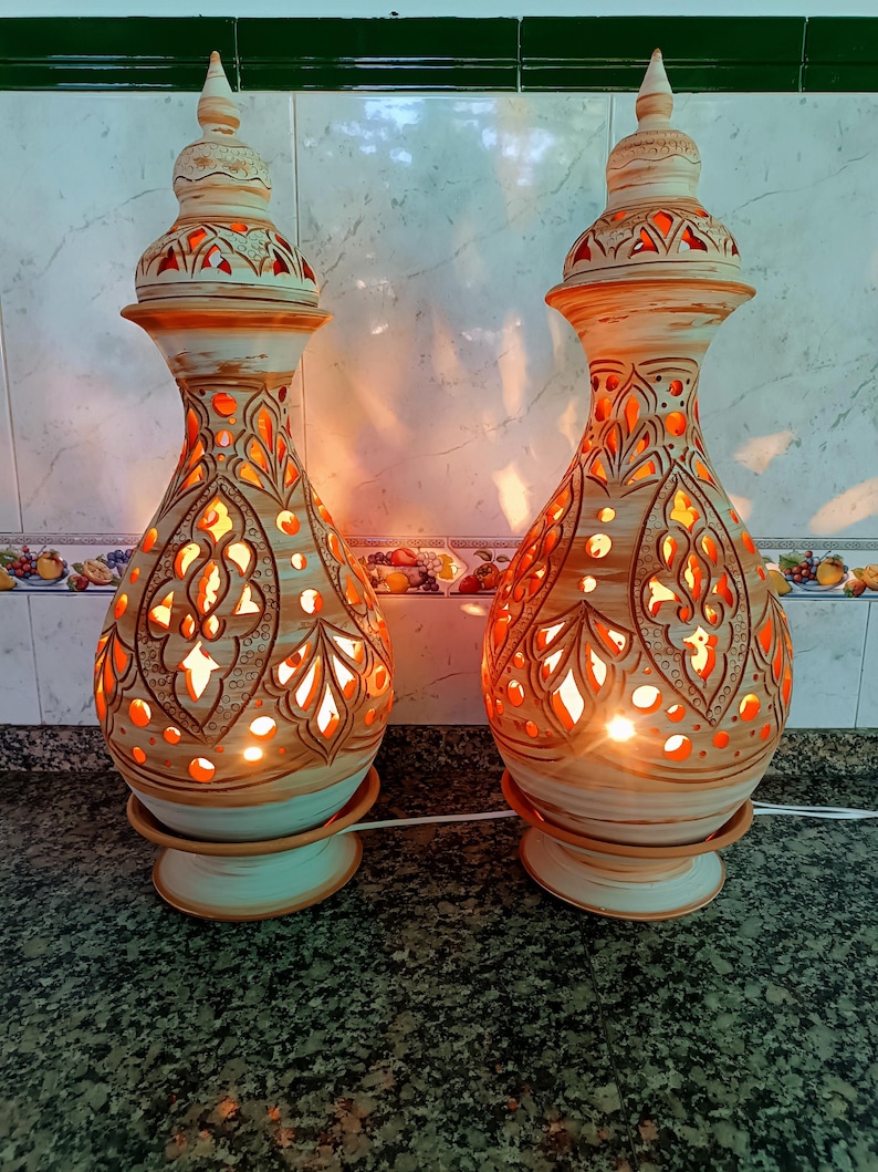 May include: Two decorative ceramic lamps with intricate cut-out patterns, emitting a warm orange glow. The lamps have a beige and brown color scheme with a unique, ornate design. Each lamp is approximately 12 inches tall.